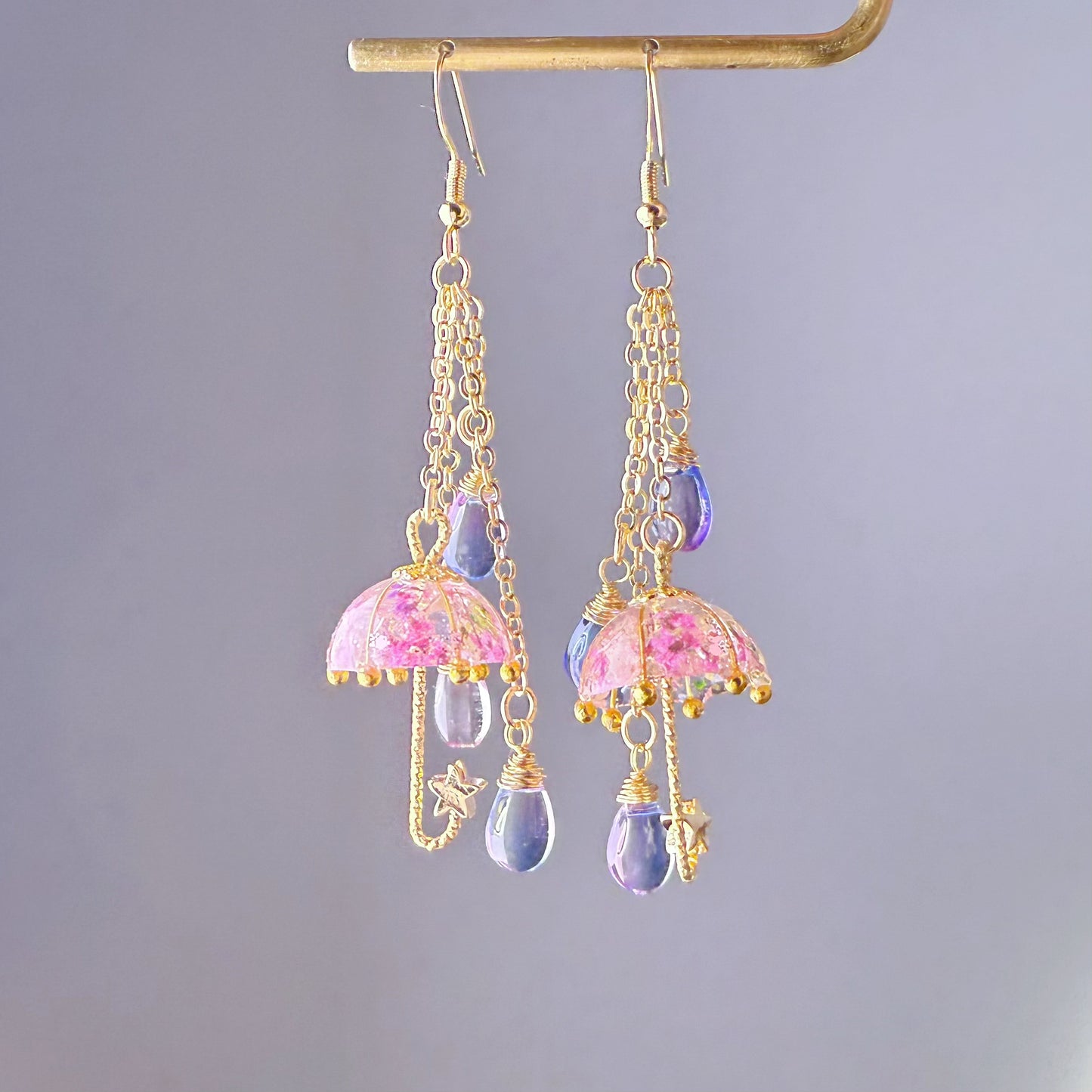 Pair of earrings with pink umbrella design and purple teardrop beads on a gray background