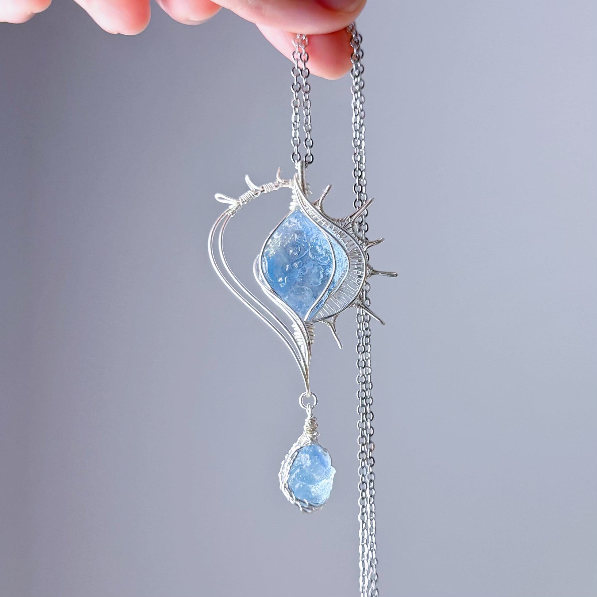 Silver necklace with a blue pendant held by a hand against a gray background