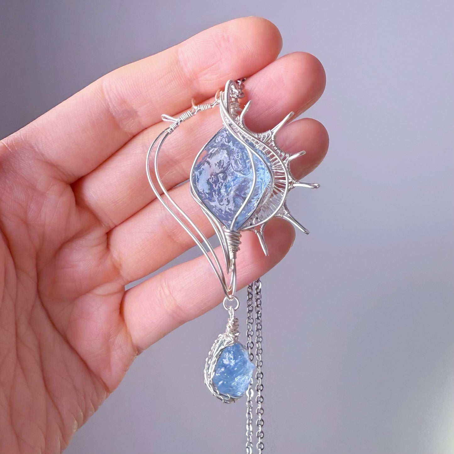 Hand holding a silver pendant with blue gemstones against a gray background