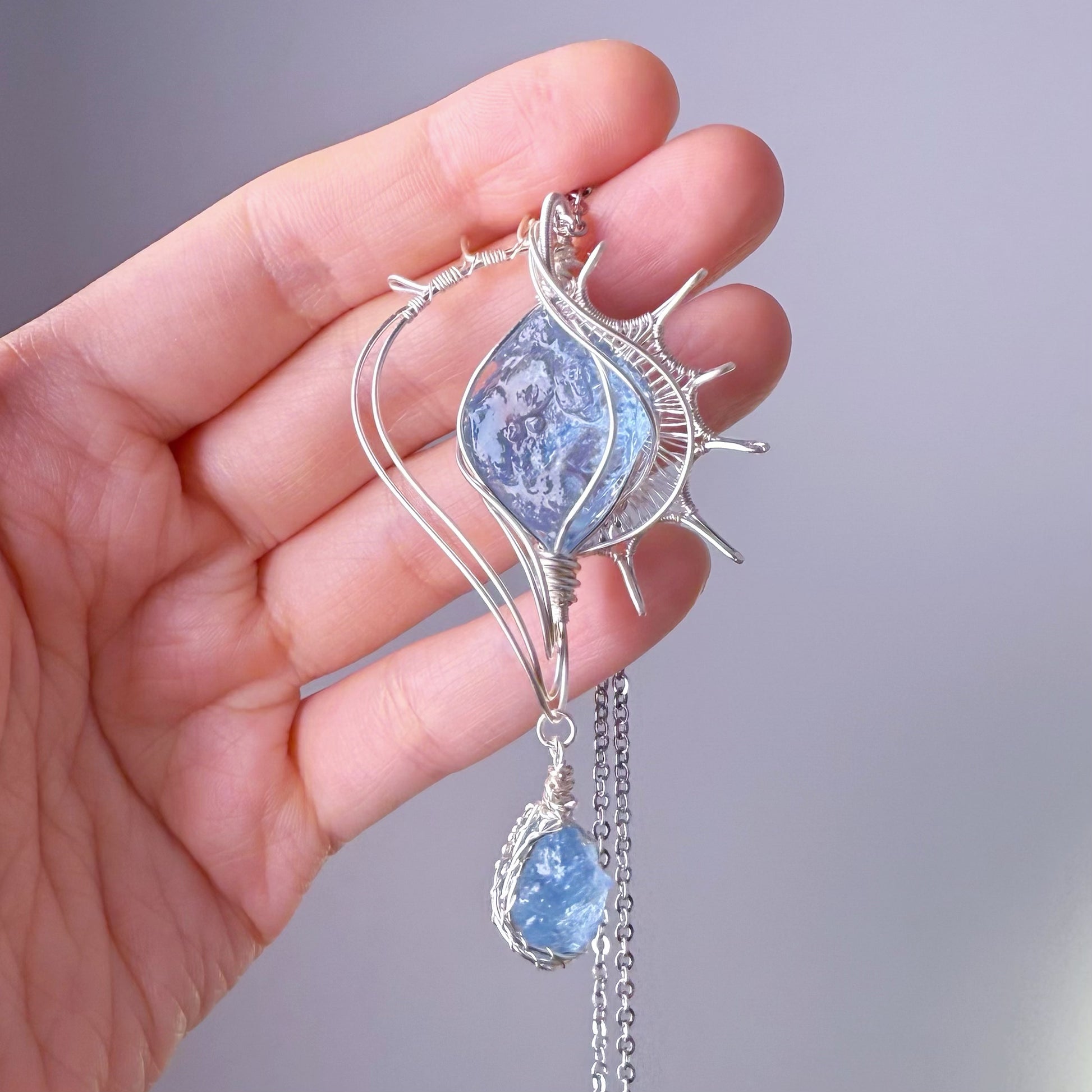 Hand holding a silver pendant with blue gemstones against a gray background