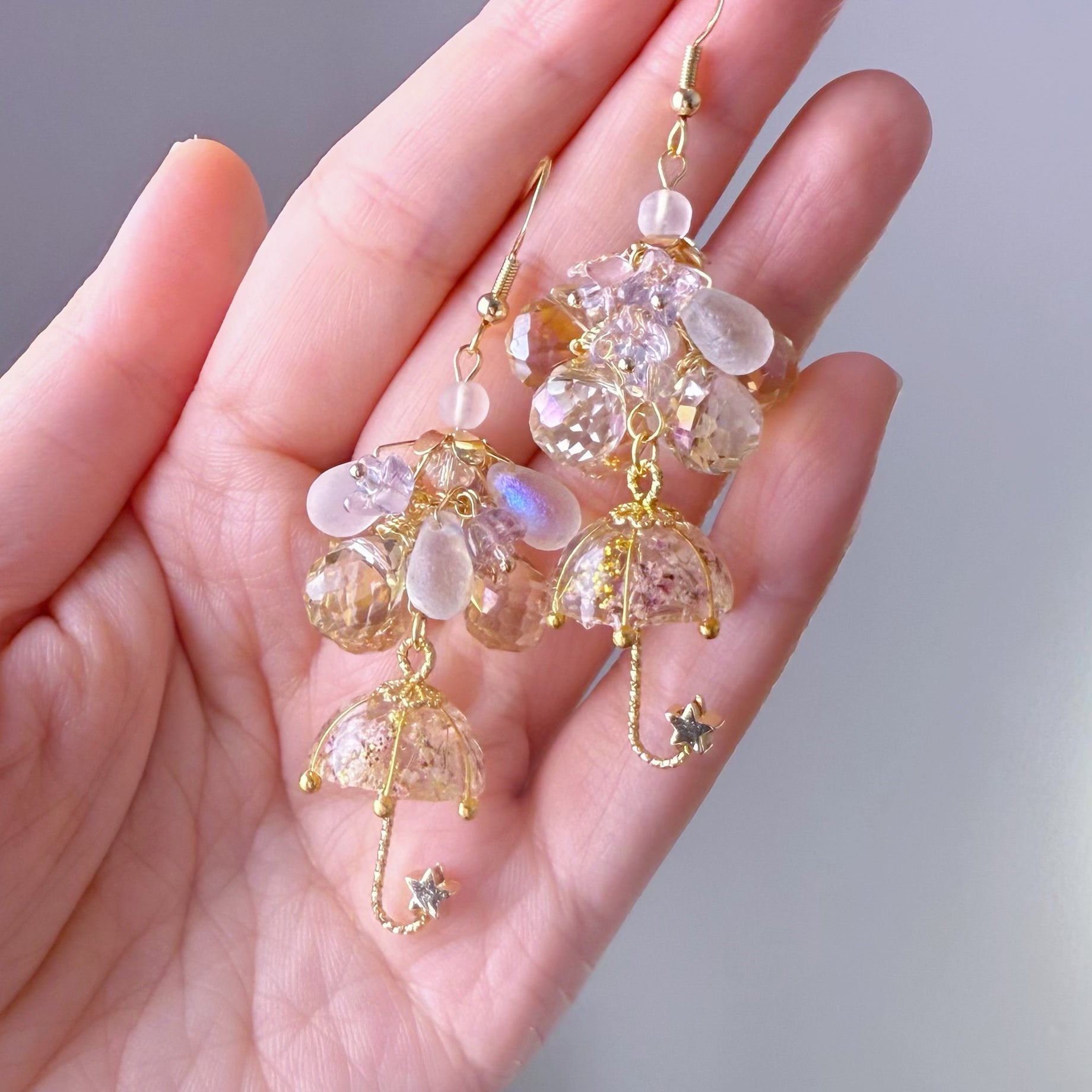Delicate earrings with crystal-like beads and gold accents held in a hand against a neutral background